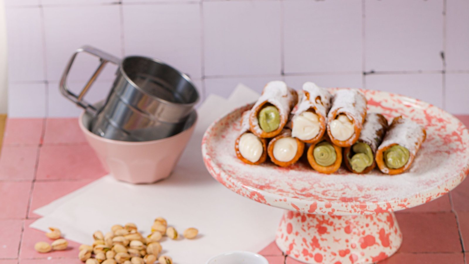 Sizilianisches Frühstück: eine Etagere voller frisch gefüllter Cannoli mit Pistazie und Ricotta, daneben ein Mehlsieb und Pistazien auf rosa Fliesen